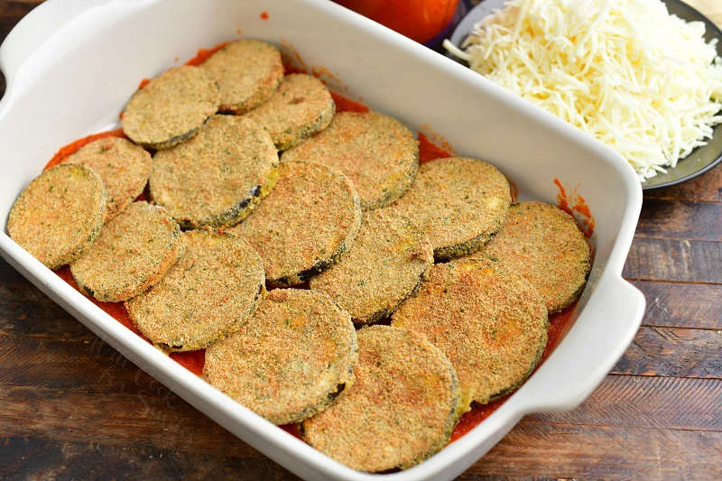 first layer of baked eggplant slices over some marinara sauce in a casserole dish, a plate full of shredded Mozzarella next to it and part of blender with marinara sauce visible