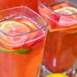 2 square glasses filled with homemade strawberry lemon lime lemonade on a wooden cutting board with lemons and limes scattered around base of glasses