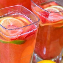 2 square glasses filled with homemade strawberry lemon lime lemonade on a wooden cutting board with lemons and limes scattered around base of glasses