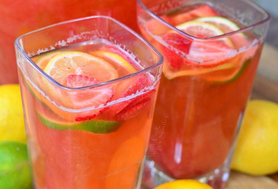 Strawberry Lemon Lime Lemonade in a clear glass with slices lemons, limes, and strawberries in it as garnish on a cutting board with lemons and lime around it 