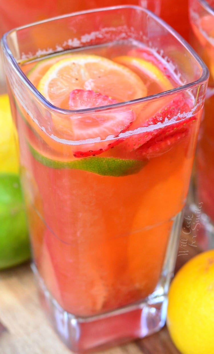 Strawberry Lemon Lime Lemonade in a clear glass with slices lemons, limes, and strawberries in it as garnish on a cutting board with lemons and lime around it 