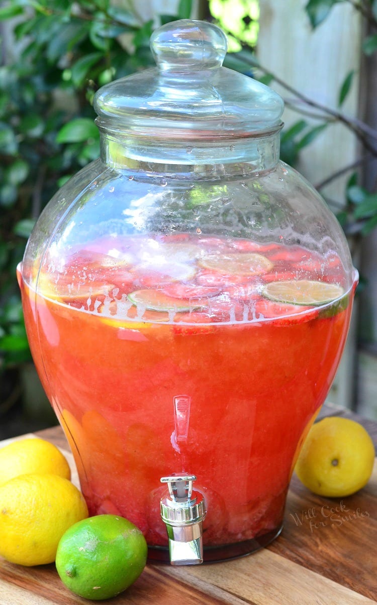 Strawberry Lemon Lime Lemonade in a clear pitcher with slices lemons, limes, and strawberries in it as garnish on a cutting board with lemons and lime around it 