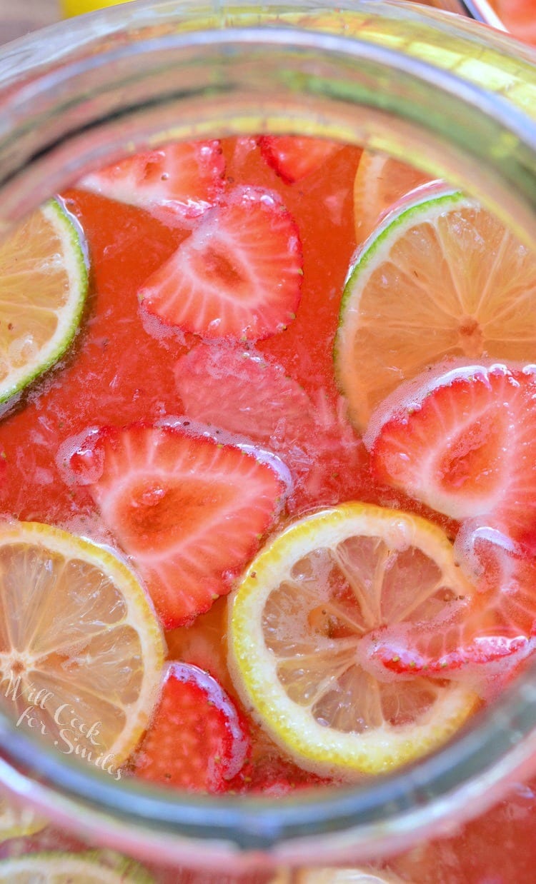 top view of Strawberry Lemon Lime Lemonade in a clear glass with slices lemons, limes, and strawberries in it as garnish on a cutting board with lemons and lime around it 