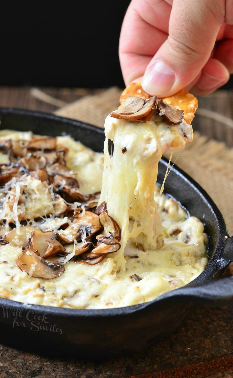 dipping a pretzel into Mushroom Leek and Gruyere Cheese Dip with mushrooms on the top in a cast iron pan 