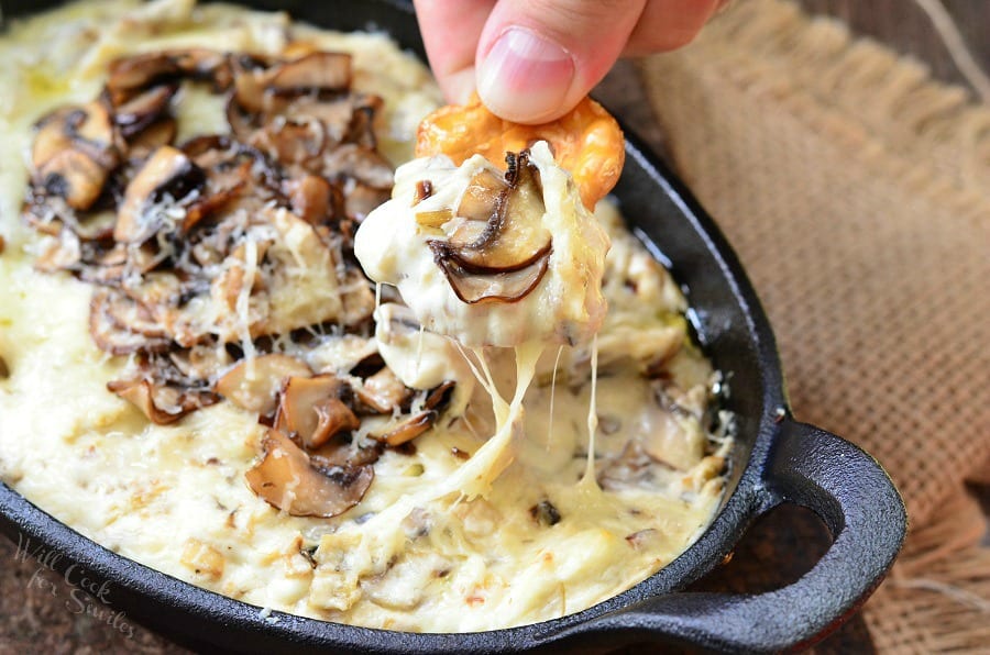 dipping a pretzel into Mushroom Leek and Gruyere Cheese Dip with mushrooms on the top in a cast iron pan 