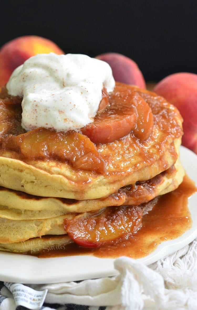 Buttermilk Pancakes with peaches on and whip cream on top and homemade syrup on a white plate with peaches in the background 