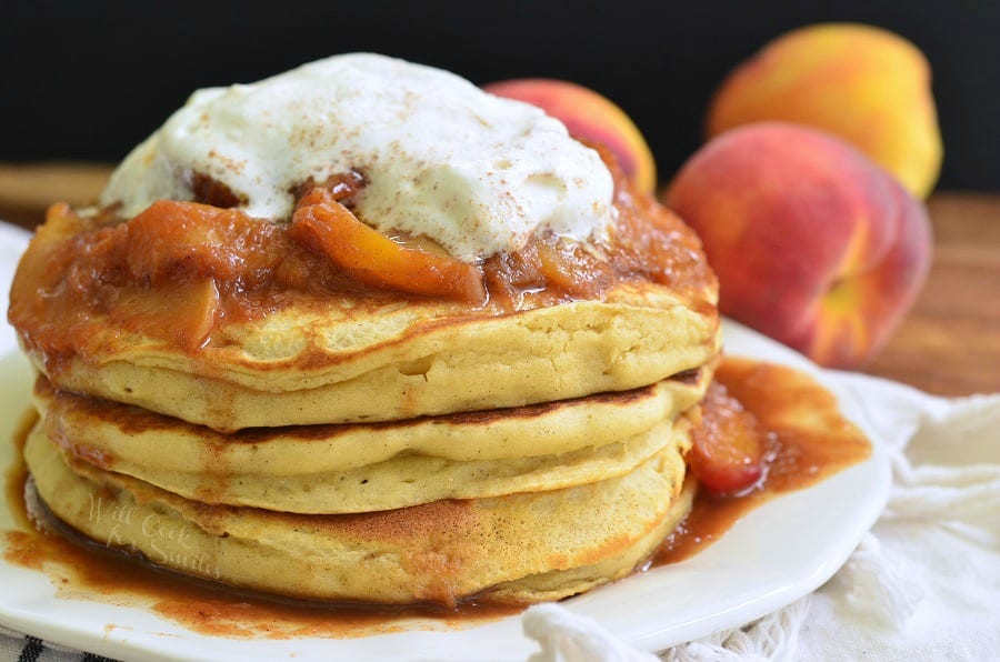 Buttermilk Pancakes with peaches on and whip cream on top and homemade syrup on a white plate and peaches in the background 