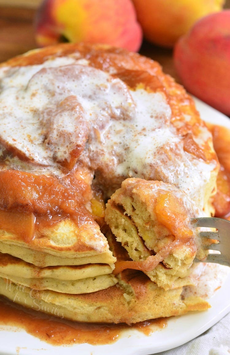 Buttermilk Pancakes with peaches on and whip cream on top and homemade syrup on a white plate with a fork lifting a bite out 