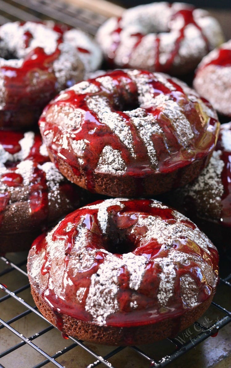 Raspberry Chocolate Doughnuts on a cooling rack with powdered sugar and raspberry sauce over the top of them on a cooling rack 