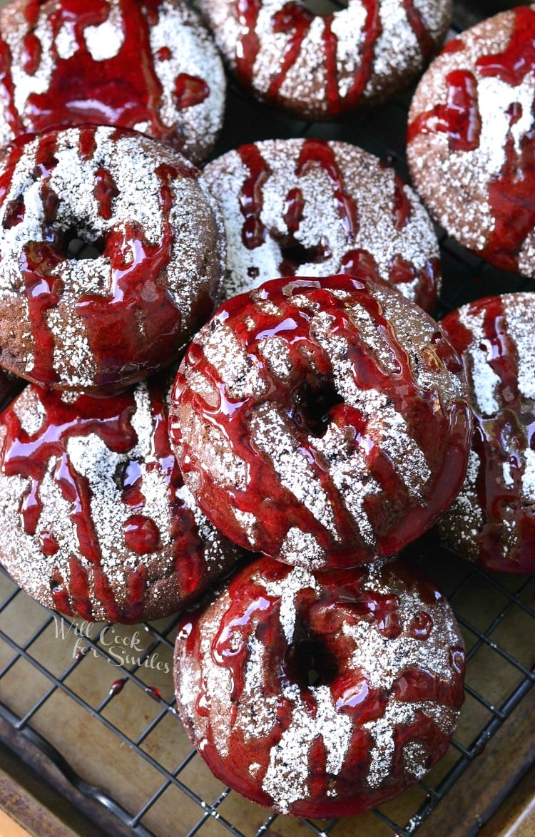 Raspberry Chocolate Doughnuts on a cooling rack with powdered sugar and raspberry sauce over the top of them on a cooling rack 