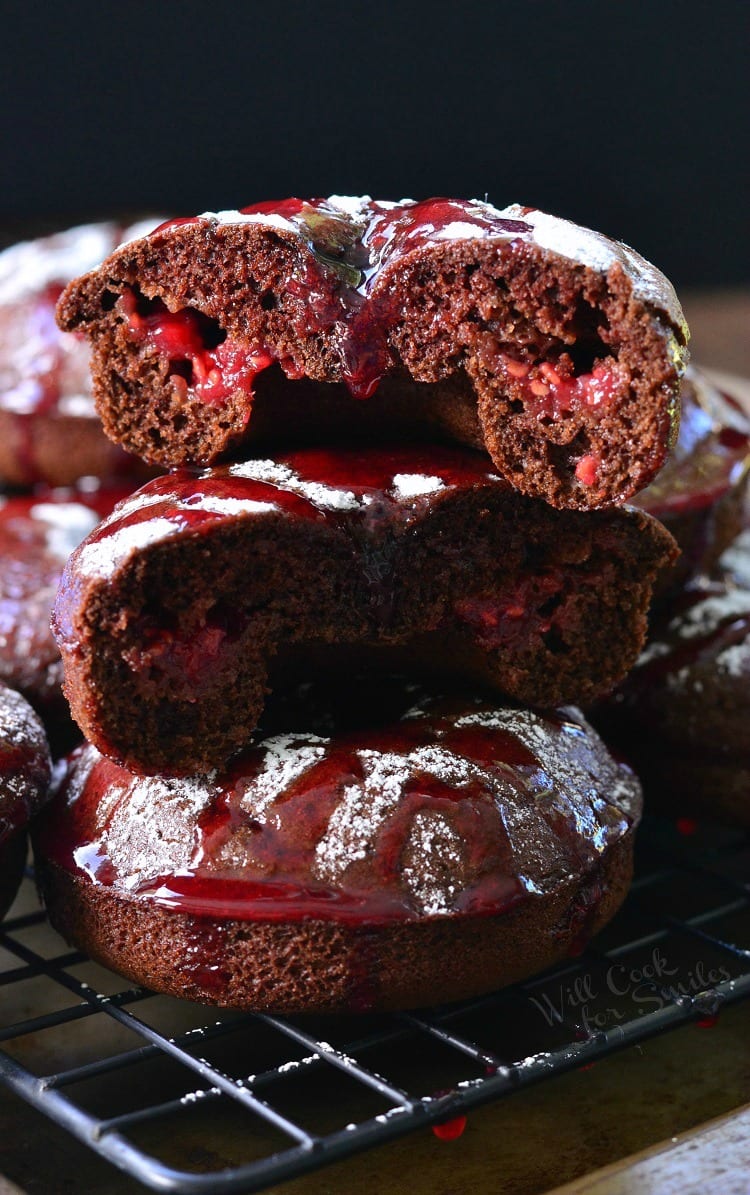 Raspberry Chocolate Doughnuts stacked up with the top one cut in half on a cooling rack with powdered sugar and raspberry sauce over the top of them on a cooling rack 