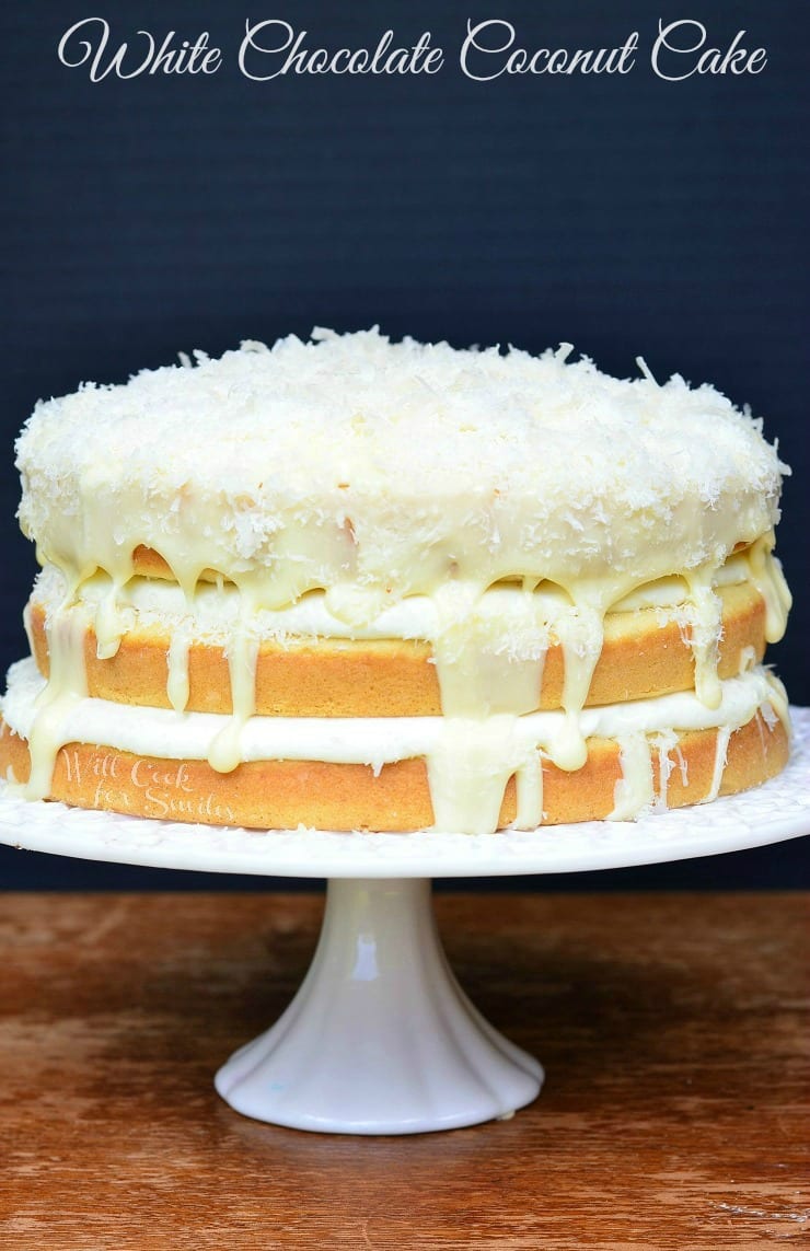 3 layers of White cake with white Chocolate and Coconut in between them and coconut on top on a white cake stand 