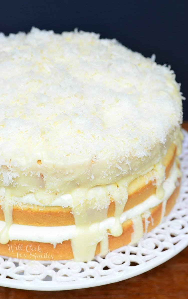 slice of 3 layers of White cake with white Chocolate and Coconut in between them and coconut on top on a piece of wax paper on a wood table with the rest of the cake in the background on the cake stand 