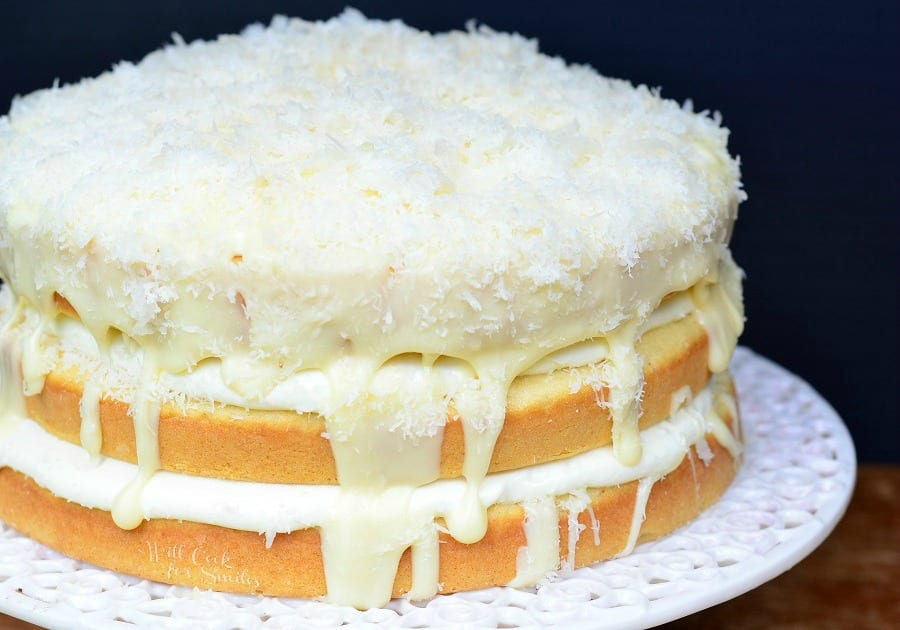 close up 3 layers of White cake with white Chocolate and Coconut in between them and coconut on top on a white cake stand 