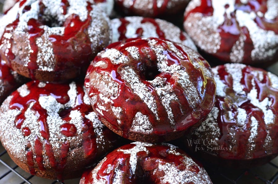 Raspberry Chocolate Doughnuts on a cooling rack with powdered sugar and raspberry sauce over the top of them on a cooling rack 