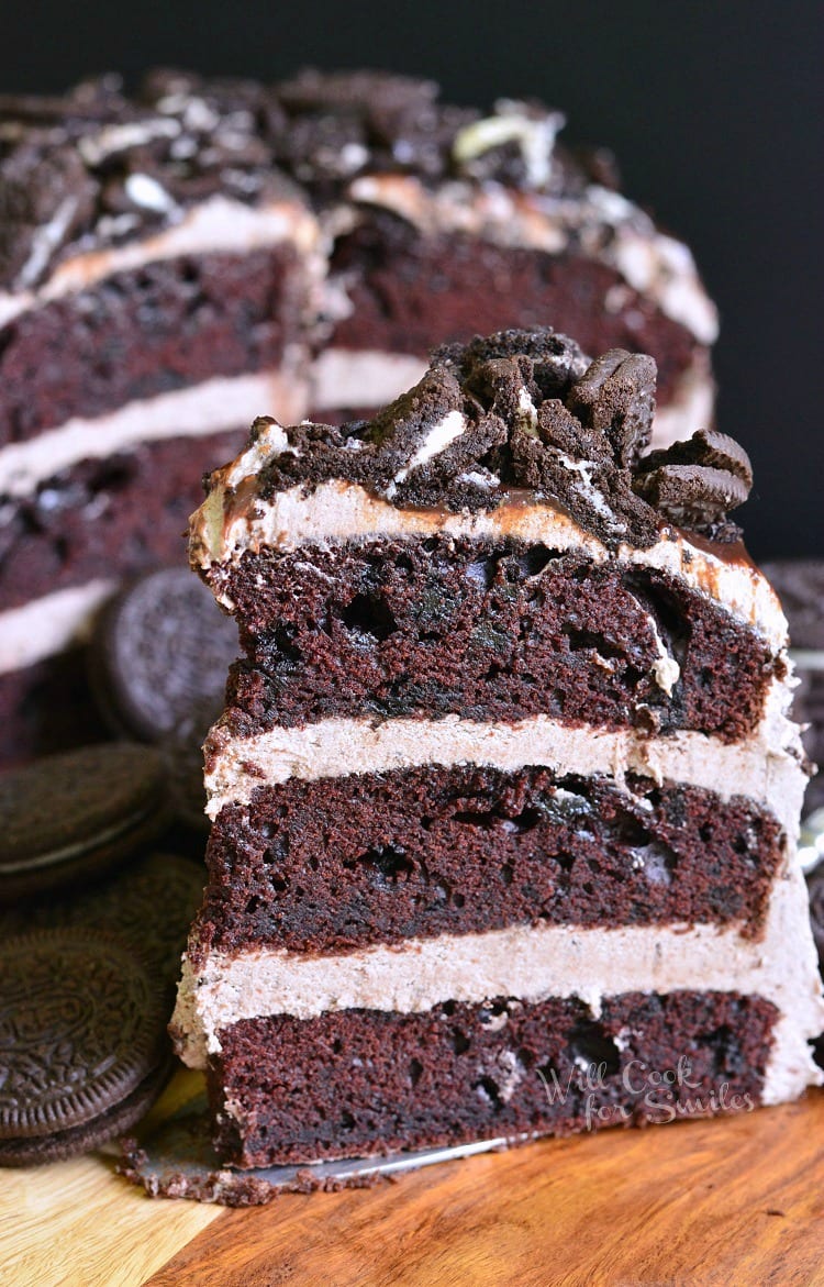 slice of Chocolate Oreo Cake on cutting board with the rest of the cake in the background 