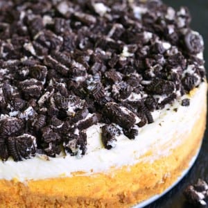 Layered Oreo Pumpkin Cheesecake with white frosting and oreo cookie crumbles on a black table.