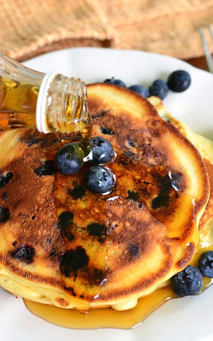 pouring maple syrup over the top of blueberry pancakes