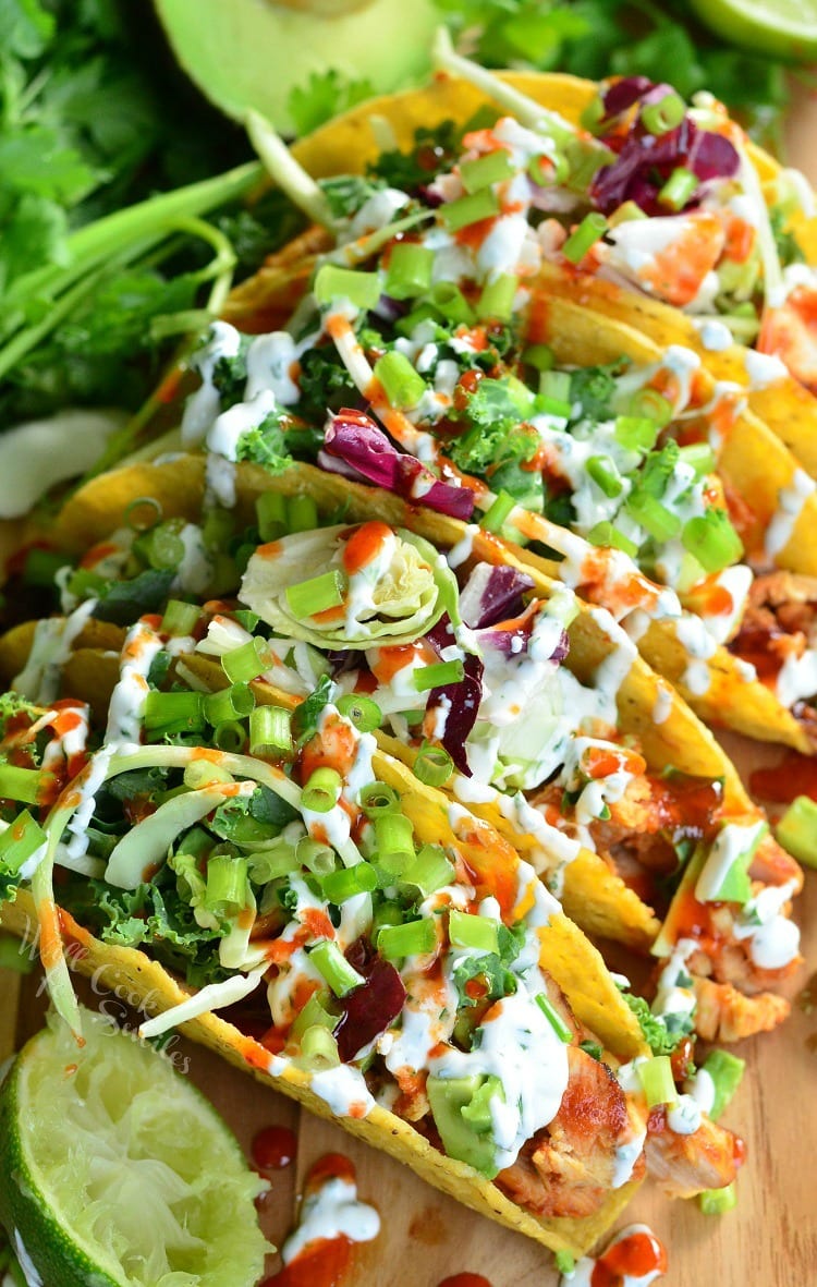 View from above of sweet and spicey siracha chicken tacos on a wooden cutting board with limes and avocado halves around them