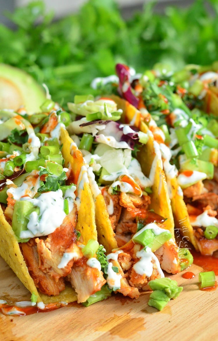 sweet and spicey siracha chicken tacos on a wooden cutting board with limes and avocado halves around them