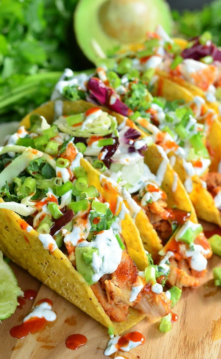 sweet and spicey siracha chicken tacos on a wooden cutting board with limes and avocado halves around them