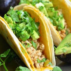 3 avocado turkey tacos on a slate table with avocados in the background and to the left of the tacos as viewed close up.