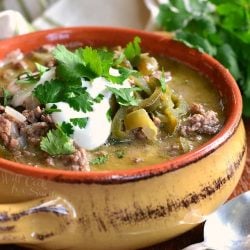 Large coup crock filled with beef and poblano enchilada soup topped with sour cream and parsley. A spoon lays at the bottom right of bowl on a wooden table.