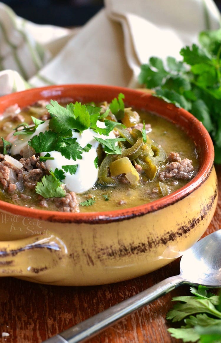 Large coup crock filled with beef and poblano enchilada soup topped with sour cream and parsley. A spoon lays at the bottom right of bowl on a wooden table.