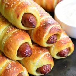Stack of homemade dijon pretzel wrapped hot dogs with maple dijon dipping sauce in a white cup to the right of the hot dogs.