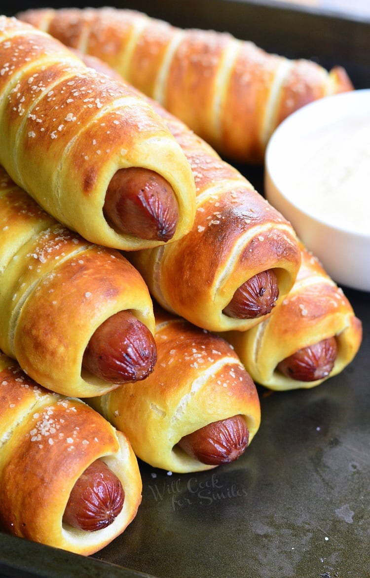 Pretzel Wrapped Hot Dogs stacked up on baking sheet with Maple Dijon Dipping Sauce in a white bowl 