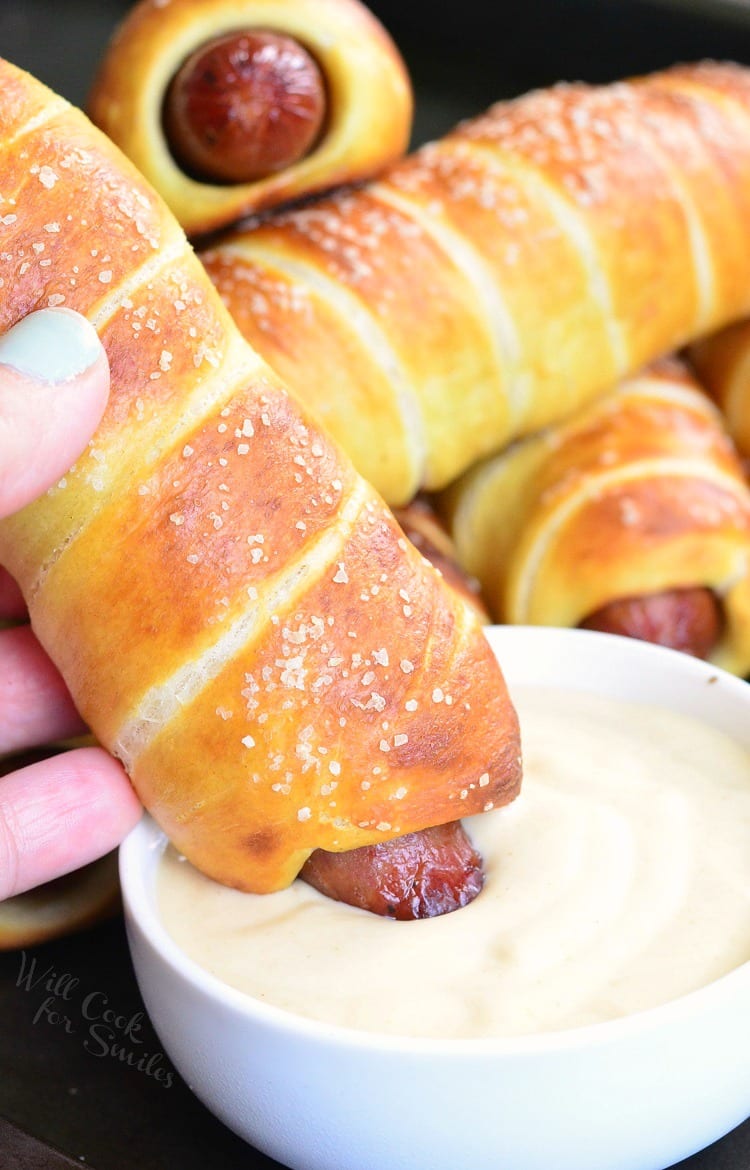 dipping Pretzel Wrapped Hot Dog into Maple Dijon Dipping Sauce in a white bowl 