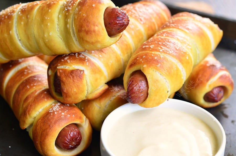 Pretzel Wrapped Hot Dogs stacked up on baking sheet with Maple Dijon Dipping Sauce in a white bowl 