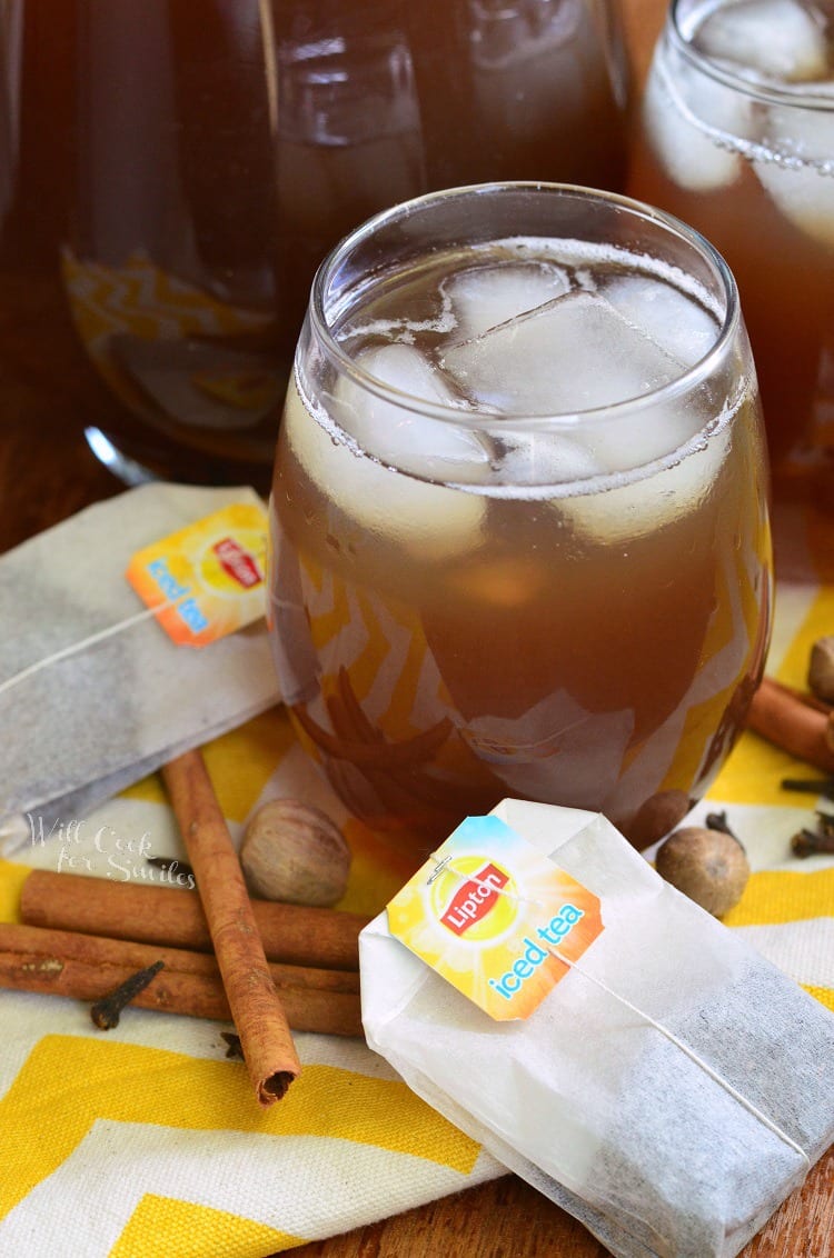 top photo tea in a glass with ice and a cinnamon  stick across the top on a yellow and white table clothe with nutmeg, clothes, and cinnamon sticks, and lipton tea bags around it   