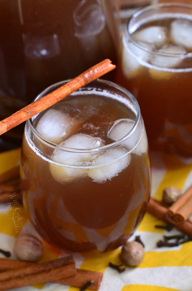 tea in a glass with ice and a cinnamon stick across the top on a yellow and white table clothe with nutmeg, clothes, and cinnamon sticks around it 