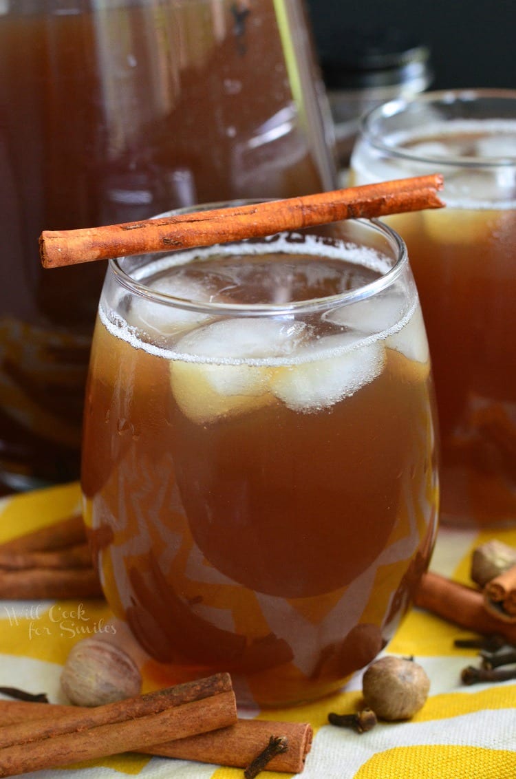 tea in a glass with ice and a cinnamon  stick across the top on a yellow and white table clothe with nutmeg, clothes, and cinnamon sticks around it   