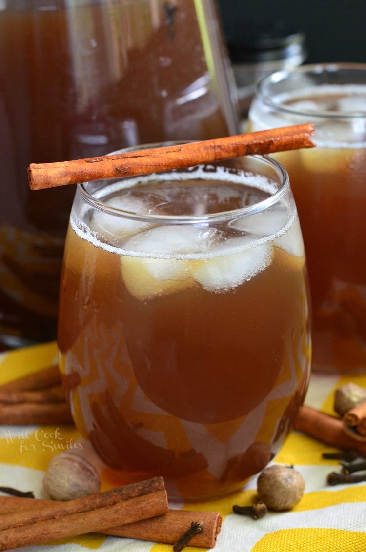 tea in a glass with ice and a cinnamon stick across the top on a yellow and white table clothe with nutmeg, clothes, and cinnamon sticks around it 