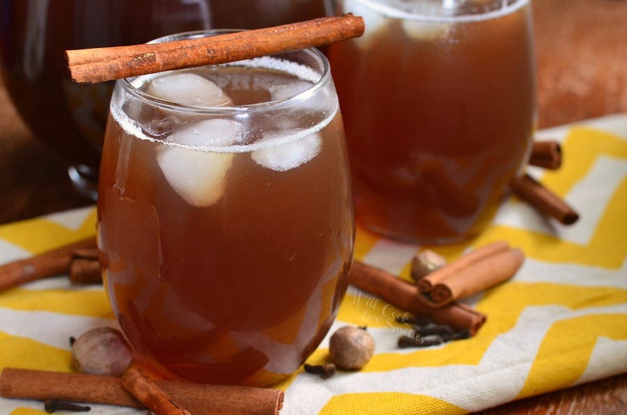 tea in a glass with ice and a cinnamon stick across the top on a yellow and white table clothe with nutmeg, clothes, and cinnamon sticks around it 