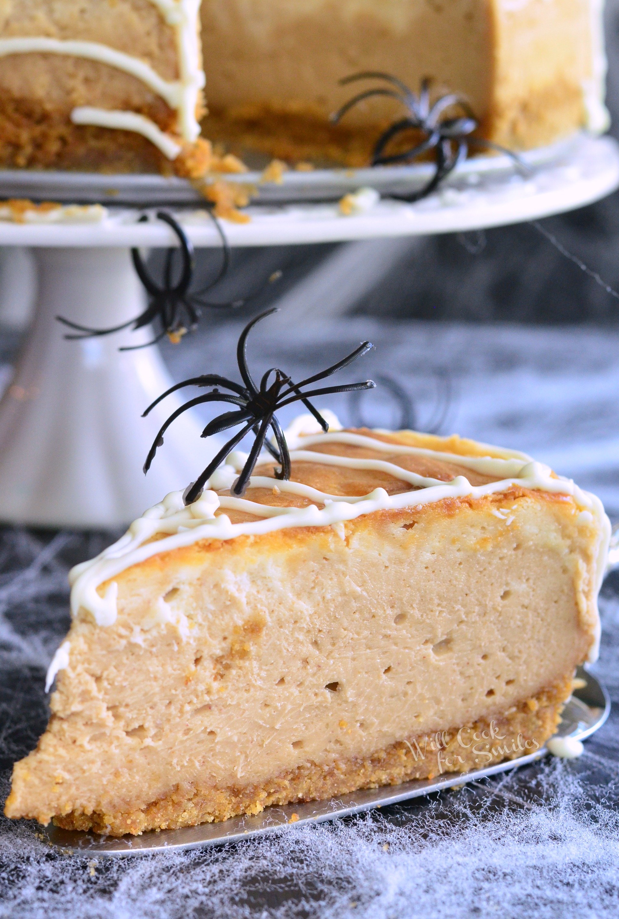 slice of cheesecake with fake spiders and the rest of the cheesecake in the background on a white cake stand 