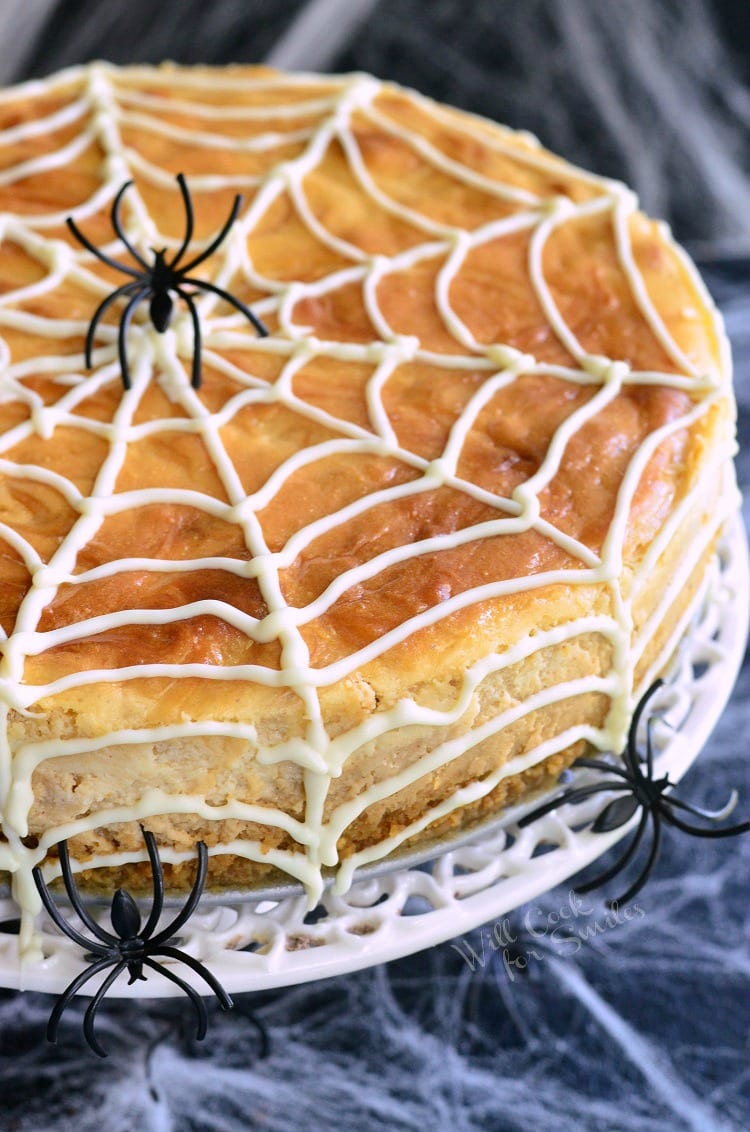 top view of cheesecake on cake stand with spider web made out of frosting on top and 3 fake plastic spiders 