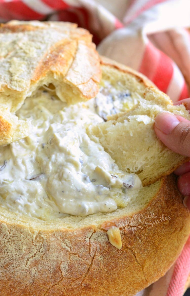 Fontina Mushroom Artichoke Dip in a bread bowl with a piece of bread being dipped inside 