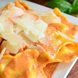 decorative white square plate filled with ravioli in creamy red pepper sauce topped with shaved parmesan.