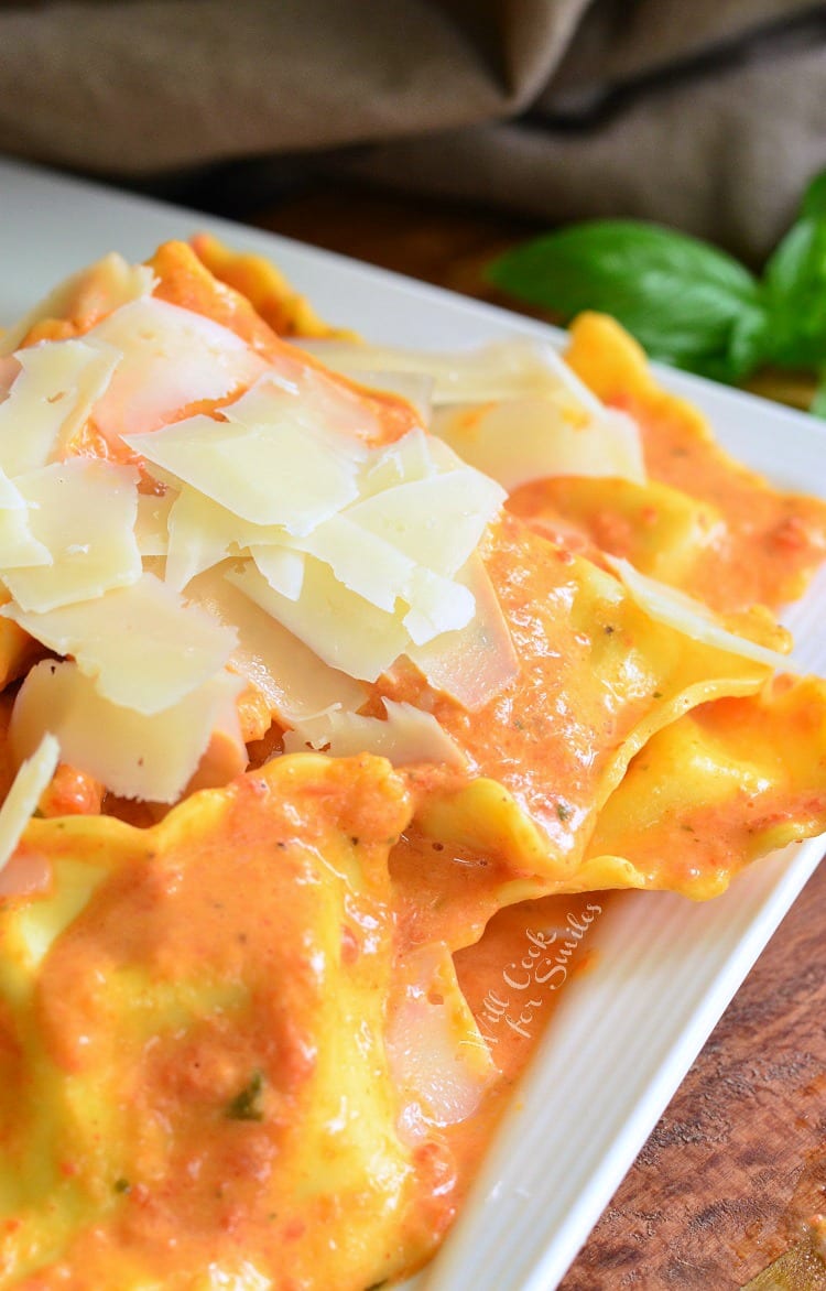 decorative white square plate filled with ravioli in creamy red pepper sauce topped with shaved parmesan.