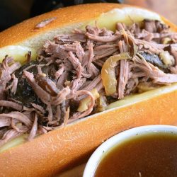 large hogie roll covered in cheese filled with slow cooker mushroom and onion italian beef on a wooden table as viewedclose up.