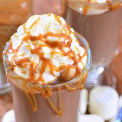 Dessert glass filled with caramel spicy hot chocolate on a wooden table with marshmellows around the base of glass as viewed from above
