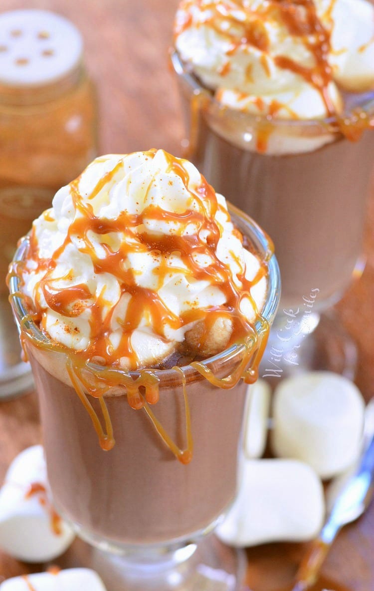 top view Caramel Spicy Hot Chocolate in a clear glass mug with whip cream and caramel on top on a cutting board with marshmallows around it 