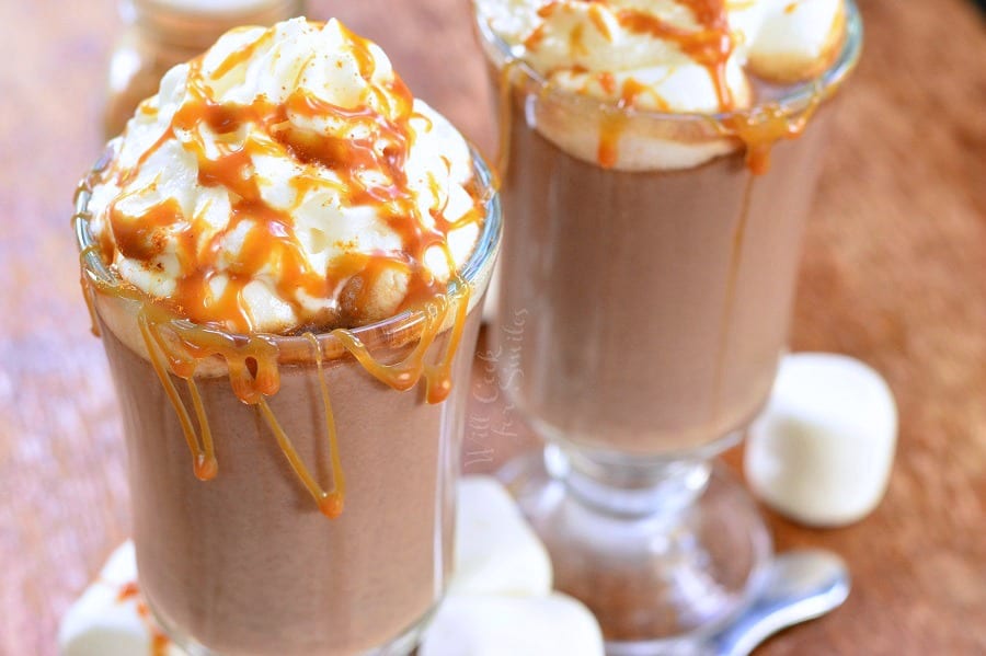 Caramel Spicy Hot Chocolate in a clear glass mug with whip cream and caramel on top on a cutting board with marshmallows around it 