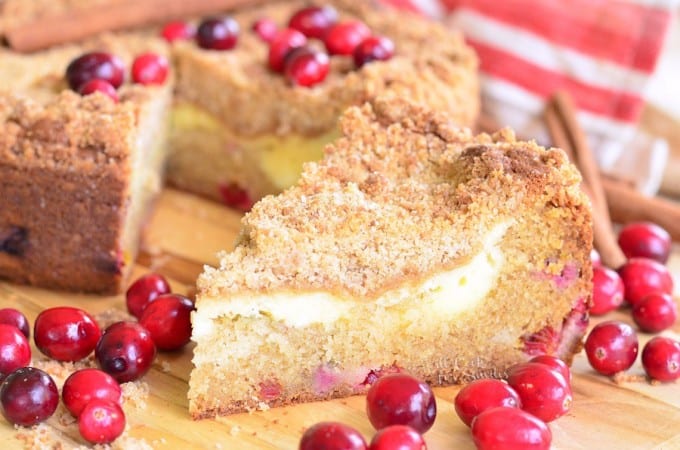 A slice of crumb-topped coffee cake with a creamy layer inside, garnished with fresh cranberries. The cake sits on a wooden board, with whole cranberries scattered around. A red and white striped cloth and cinnamon sticks are in the background.