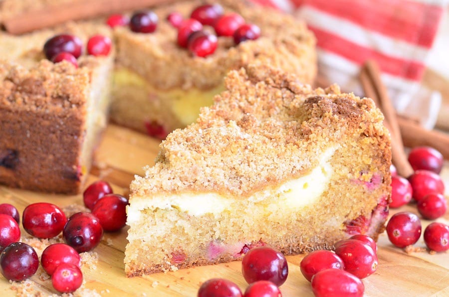 a slice of Cranberry Cream Cheese Coffee Cake surrounded by cranberries on a cutting board with the rest of the cake in the background 