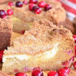 1 slice of cranberry cream cheese coffee cake on a wooden cutting board with the rest of the cake sitting behind topped with cranberries.