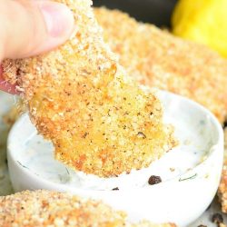 Lemon pepper baked chicken tenders around a dish of ranch with a whole lemon in the background and 1 tender being dipped into sauce.